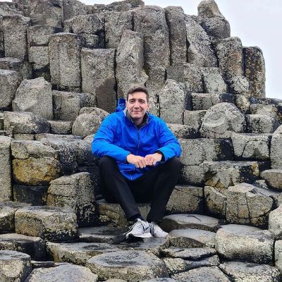 Oliver Phelps is sitting on the rock as he is wearing a blue jacket, black pants, and white sneakers.